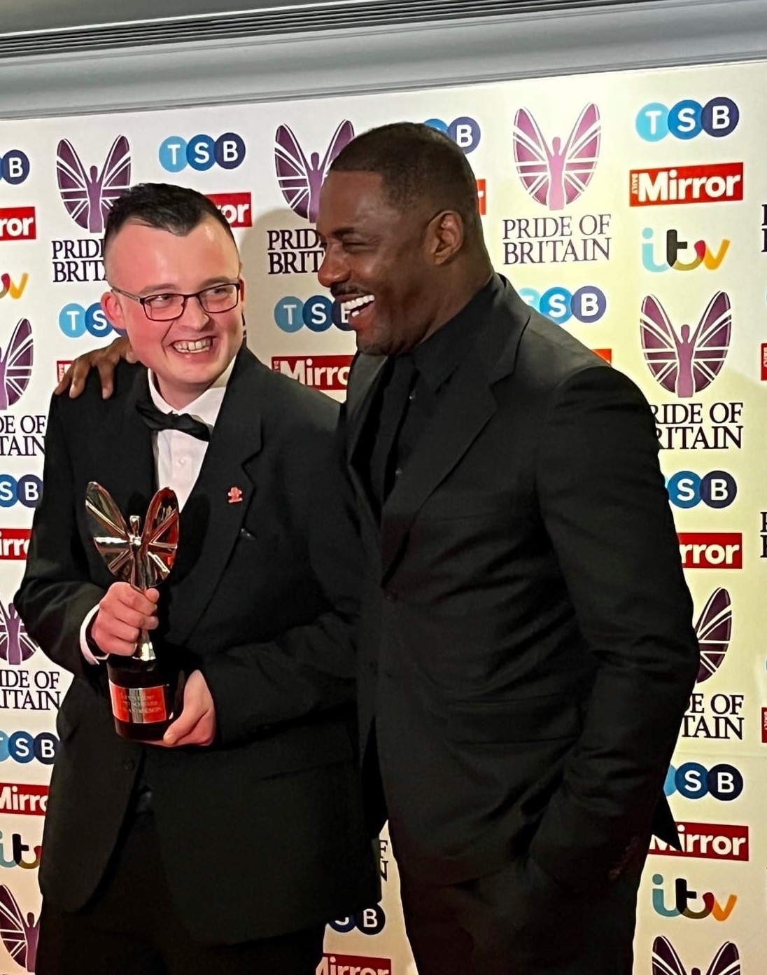 Image shows actor Idris Elba with Alex holding his award on the red carpet.